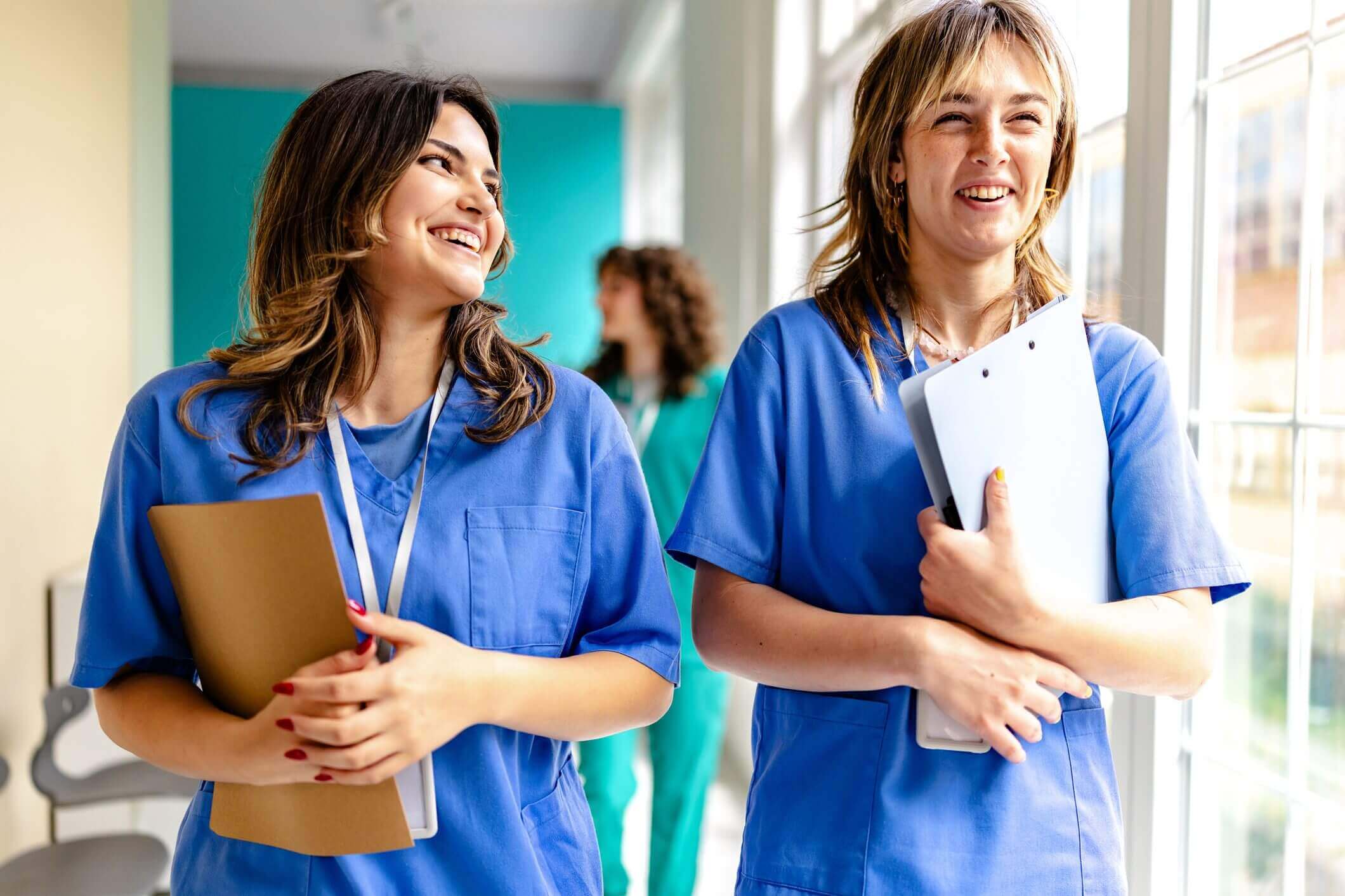 Two female healthcare workers smiling, holding folders, in a hospital setting.